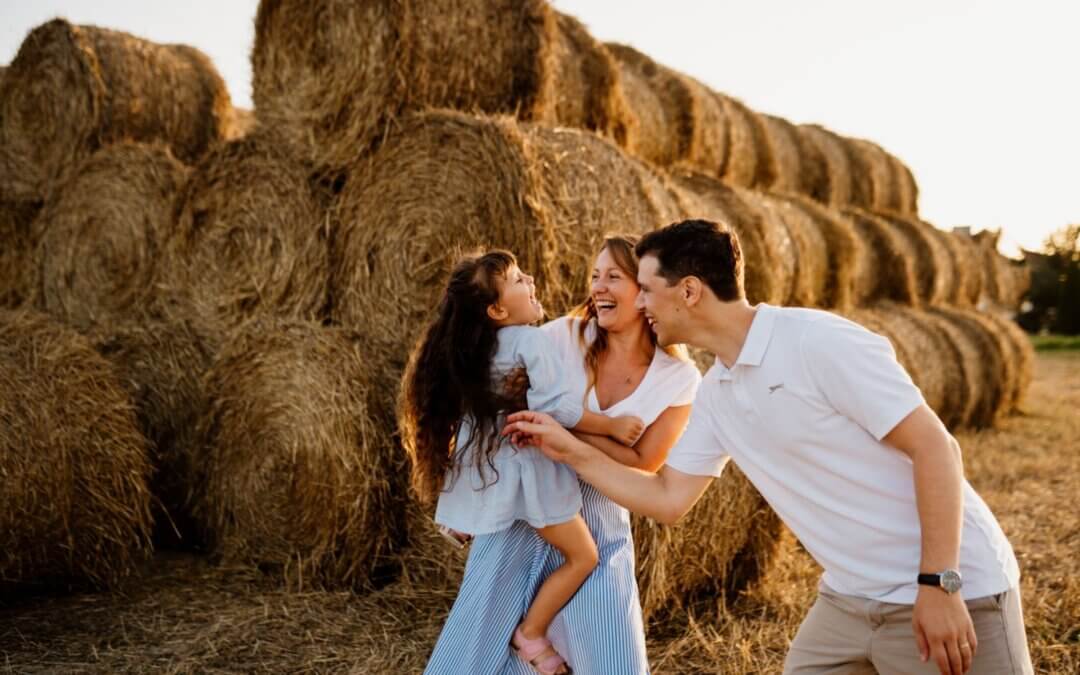Mama trzyma córkę na rękach, a tata stoi obok nich w trakcie sesji rodzinnej.